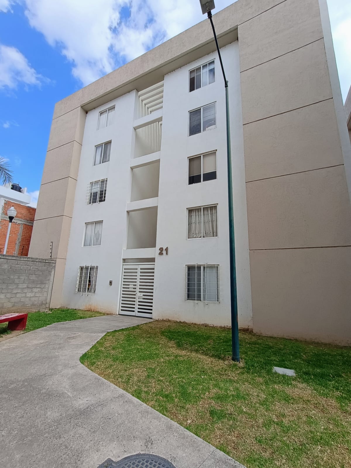 A multi-story residential building is displayed, featuring a light-colored facade. Ground-level access is seen through a gated entrance. The surrounding area includes a path bordered by grass and small shrubbery, with a glimpse of a red bench nearby.