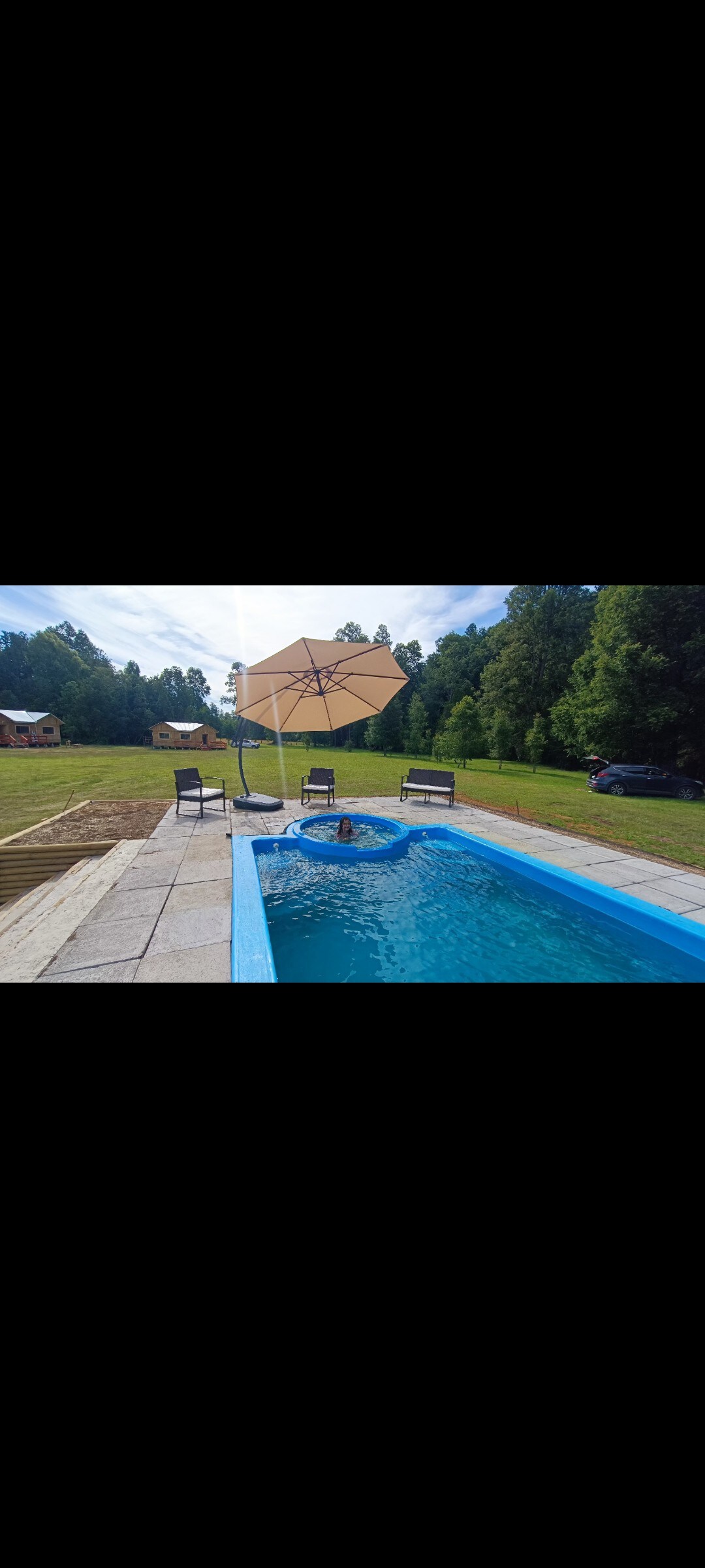 A serene outdoor area features a rectangular pool surrounded by greenery. Four chairs are arranged near the pool, complemented by a large umbrella providing shade. The sky is clear, highlighting the tranquil setting.