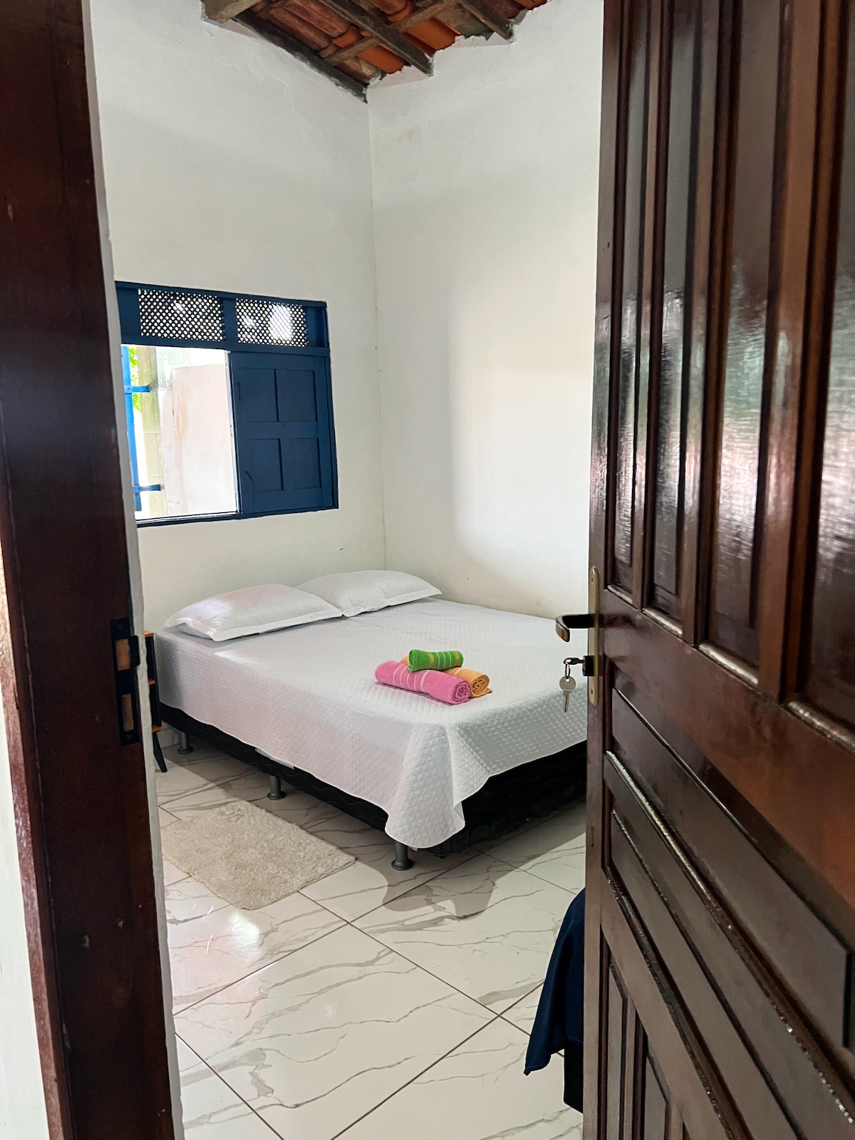 A compact bedroom is visible through an open door, featuring a spacious bed covered with a white bedspread. A mirror reflects natural light from a window with blue frames, while colorful towels in shades of pink and green are neatly arranged on the bed.
