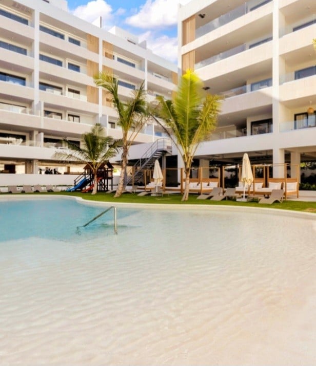 A spacious pool area is surrounded by lush palm trees and sun loungers. The well-maintained swimming pool reflects clear blue skies, while a two-story building provides a backdrop with several balconies overlooking the pool. Umbrellas are positioned near the loungers for shade.