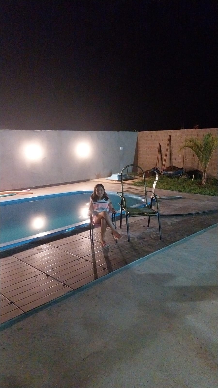 An outdoor area features a serene swimming pool illuminated by soft lights along the surrounding walls. A child sits on a chair beside the pool, surrounded by concrete and wooden decking, with grass and a small palm plant visible in the background.