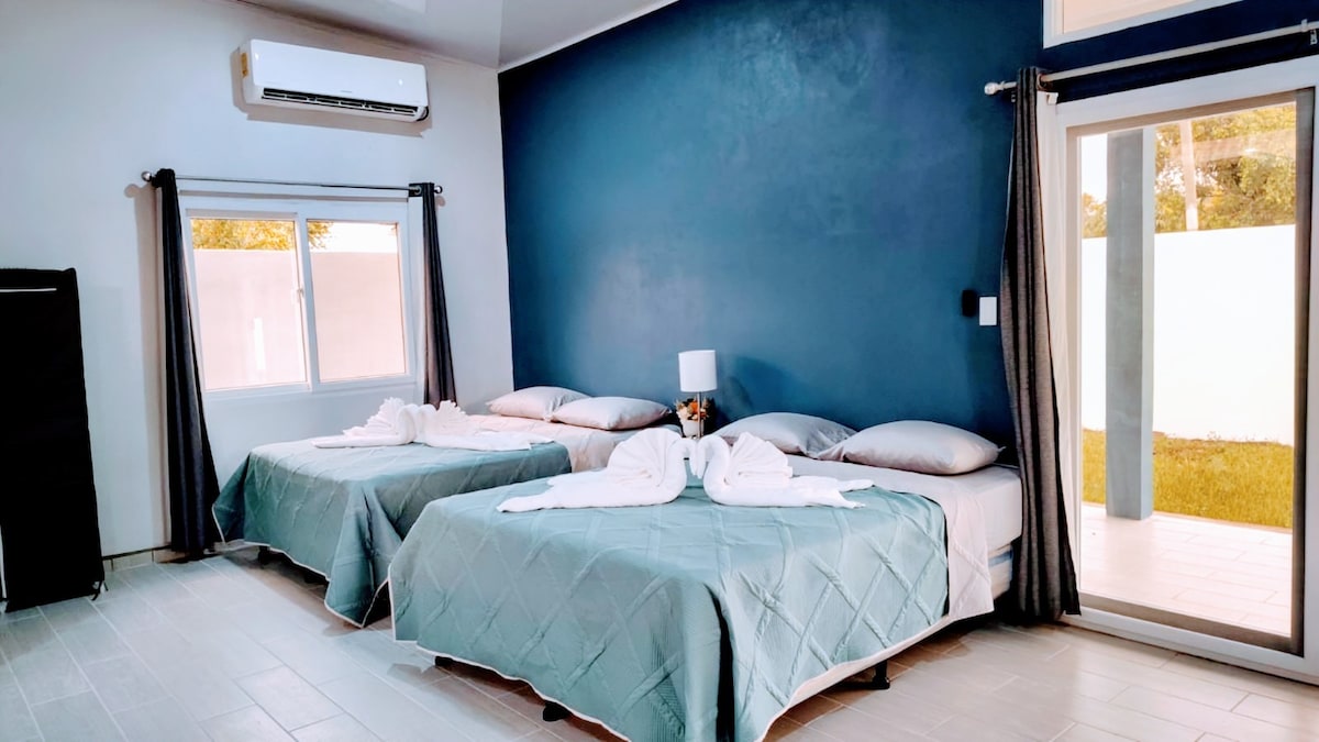 A spacious sleeping area is presented with two neatly made beds adorned in soft, muted linens. Natural light streams through a large window beside the outdoor access. A small lamp is positioned on a bedside table, and air conditioning is visible on the wall.