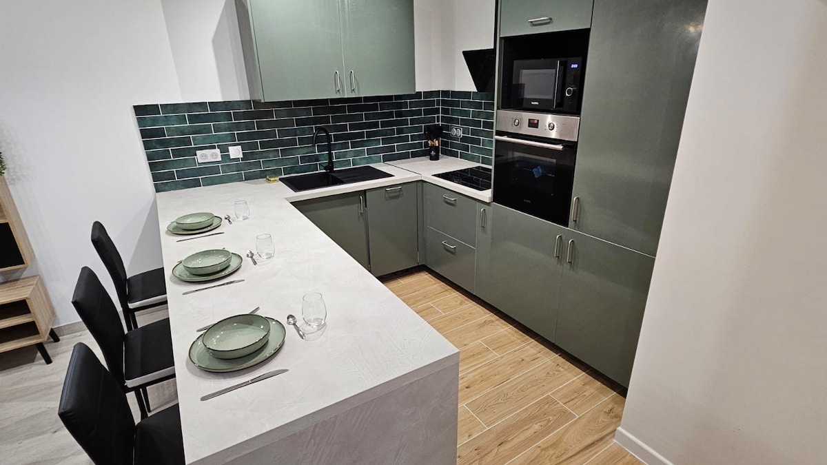 The modern kitchen features sleek cabinets in a muted green finish. A spacious countertop with seating for four is accompanied by dining ware set. Appliances include a built-in oven, microwave, and cooktop, with a stylish backsplash adding a polished touch.