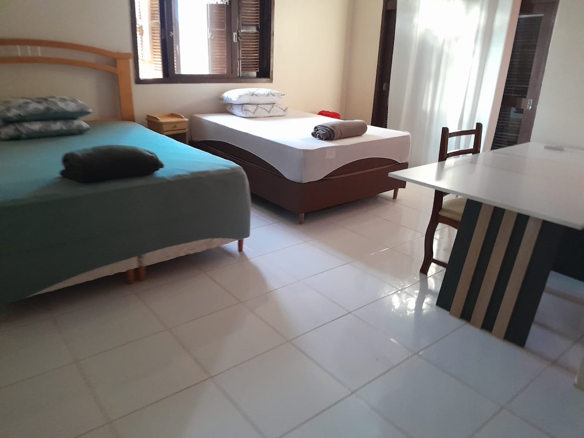 The room includes two spacious beds, each adorned with bedding in complementary tones. Natural light filters through multiple windows, and a simple dining table with a striped design occupies one corner, enhancing the functional layout of the space.