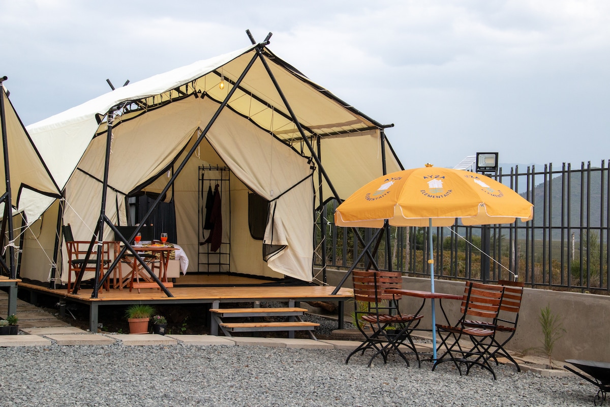 A tented structure with an open front showcases a dining area set with a table and chairs. An orange umbrella provides shade over a small outdoor seating area. Gravel paths and potted plants surround the entrance, enhancing the natural setting.