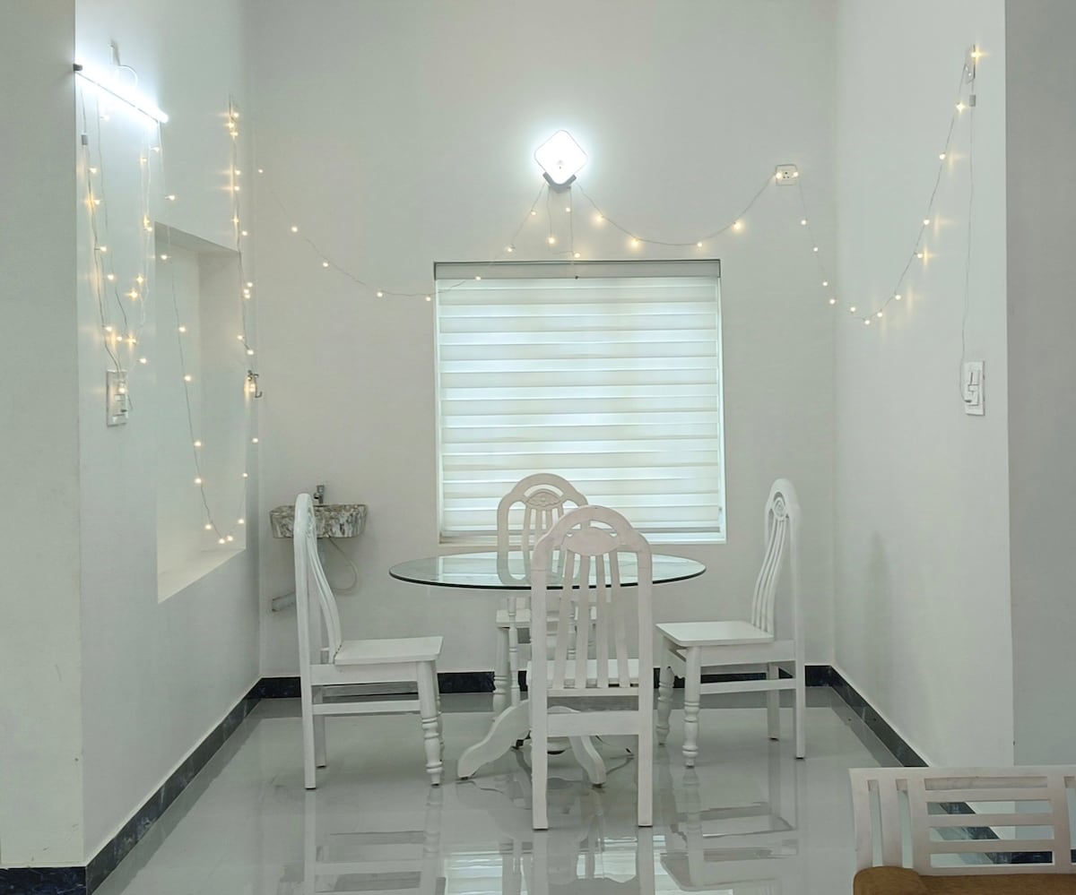 A light-filled dining area features a round glass table surrounded by four white chairs. Soft lighting is provided by string lights adorning the walls, enhancing the bright and spacious feel of the space. A window with sheer blinds allows natural light to enter.