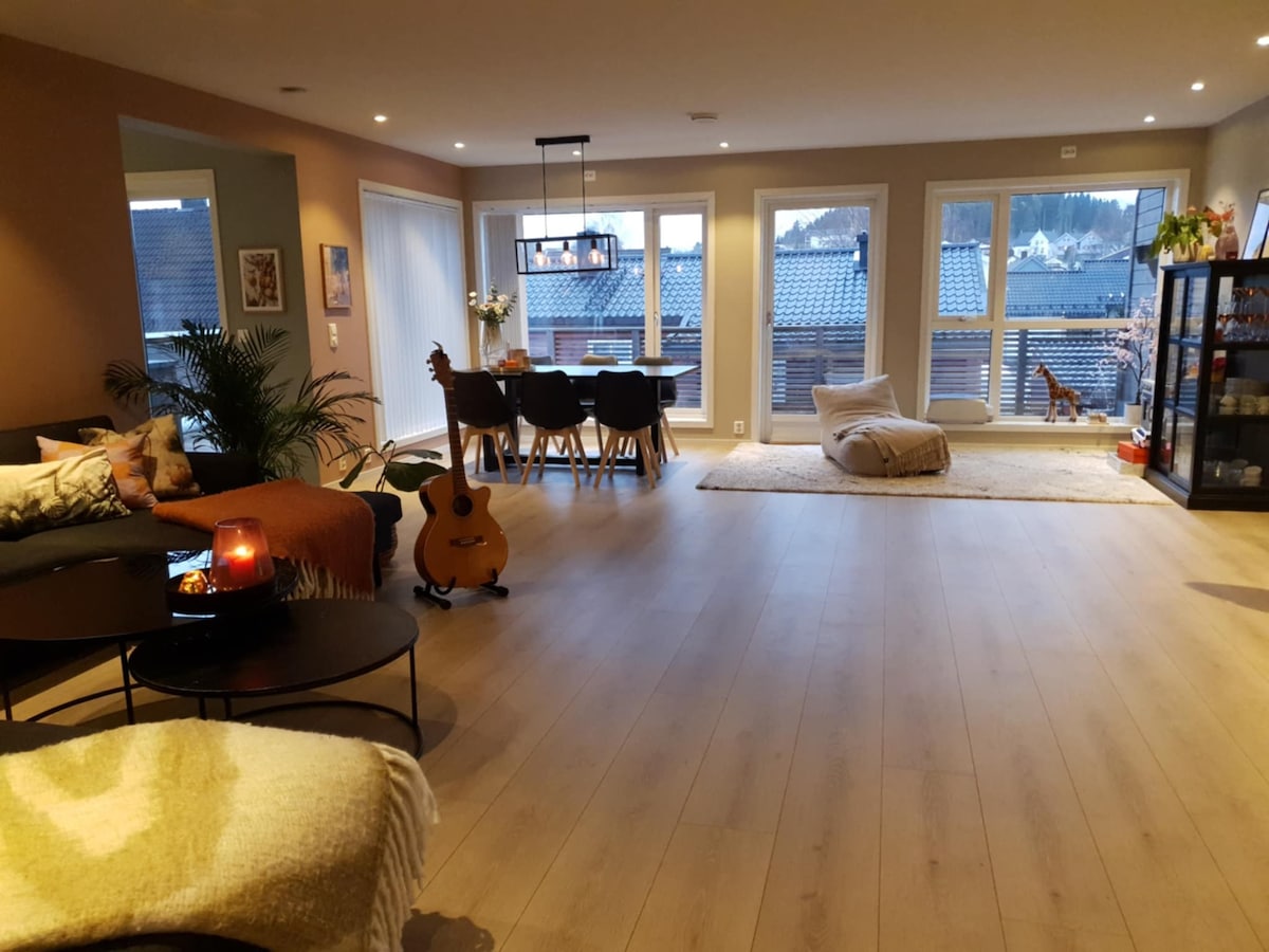 An open living space features large windows that allow natural light to fill the room. A cozy seating area is arranged with a couch and decorative throw blankets. A dining table is set for six, while plants add greenery. An acoustic guitar rests in the foreground.