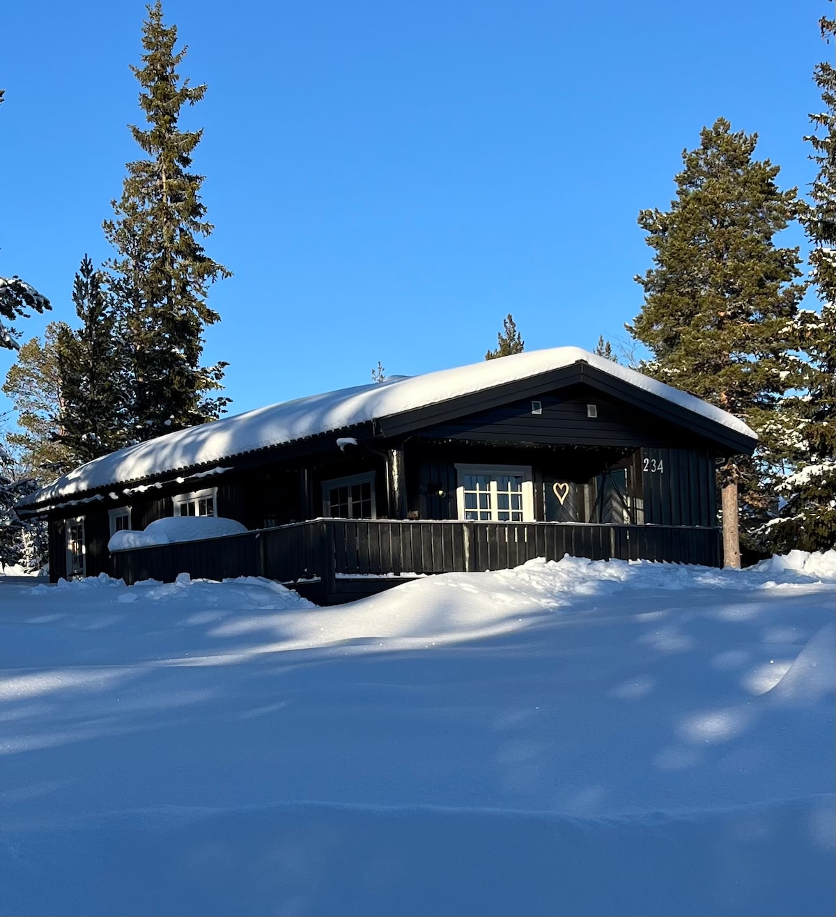 A charming cabin is nestled among tall evergreen trees, covered in a blanket of fresh snow. The structure features a sloped roof and a large front porch that overlooks the winter landscape, with sunlight illuminating the scene under a clear blue sky.