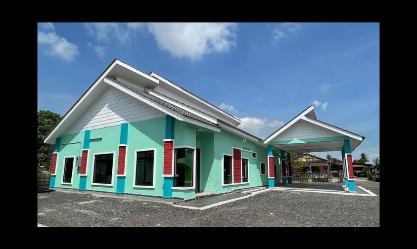 A modern, single-story villa is depicted in the image, showcasing a light green exterior accented with blue and red detailing. Multiple windows allow natural light to enter, reflecting a spacious layout. A gravel driveway leads to a covered entrance, surrounded by a clear blue sky.