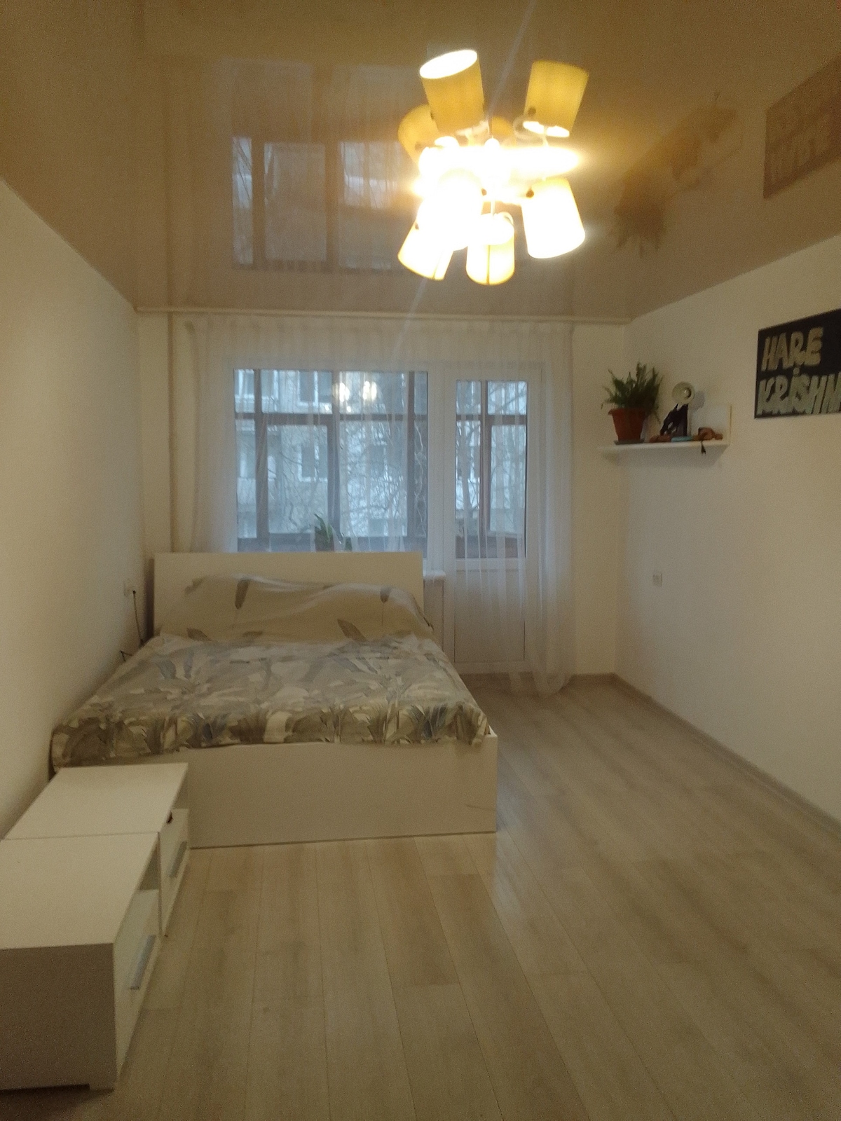 A comfortable sleeping area features a spacious bed with a light-colored patterned duvet. Warm light is provided by a unique ceiling fixture. Large windows allow natural light to illuminate the room, with sheer curtains softening the view. A small shelf holds decorative items and plants.