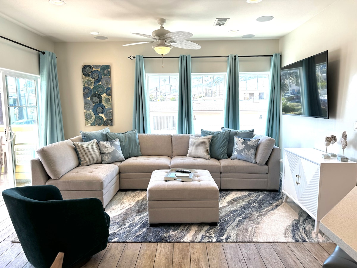 A spacious living area features a large sectional sofa adorned with blue and gray cushions. Natural light streams in through multiple windows, complemented by light blue curtains. A flat-screen TV is mounted on the wall opposite the seating, with a stylish area rug enhancing the floor.