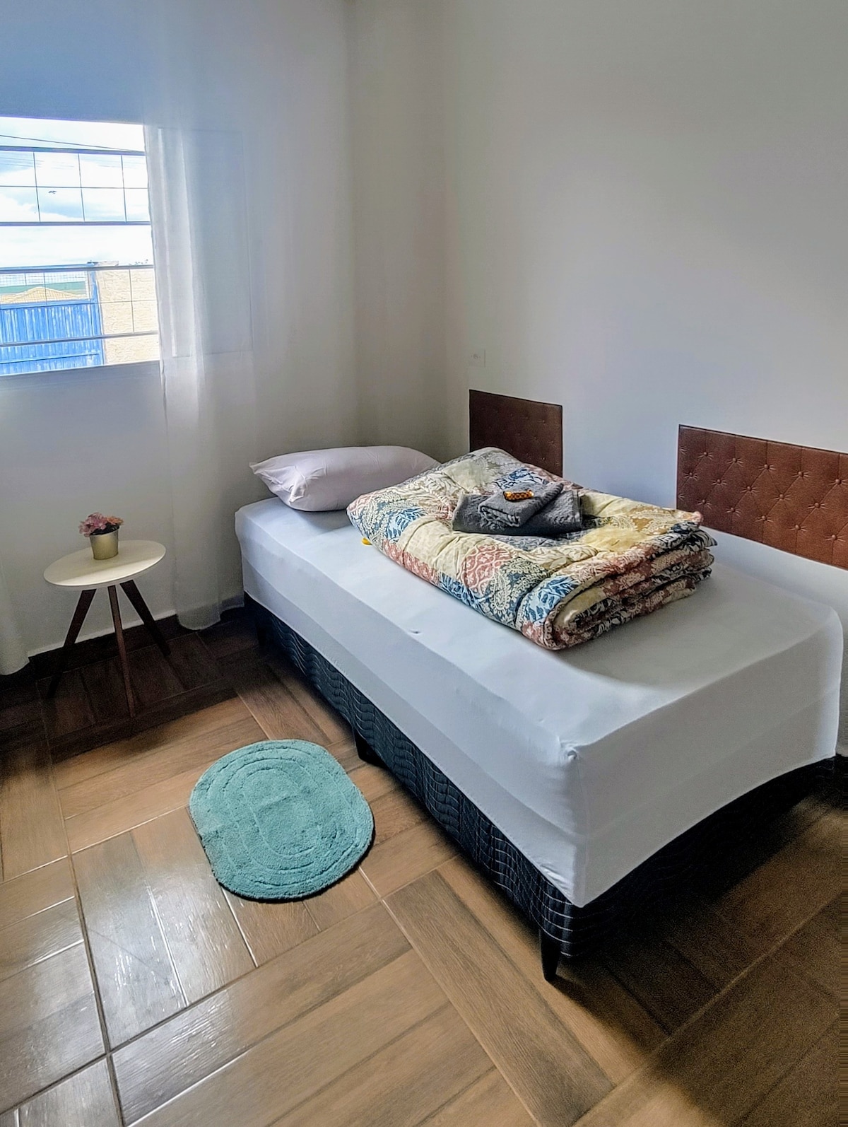 A bright bedroom features a spacious single bed layered with a colorful blanket and a decorative pillow. A small round side table holds a vase with a flower, while a soft area rug is placed on the wooden floor. Natural light streams in from the window.