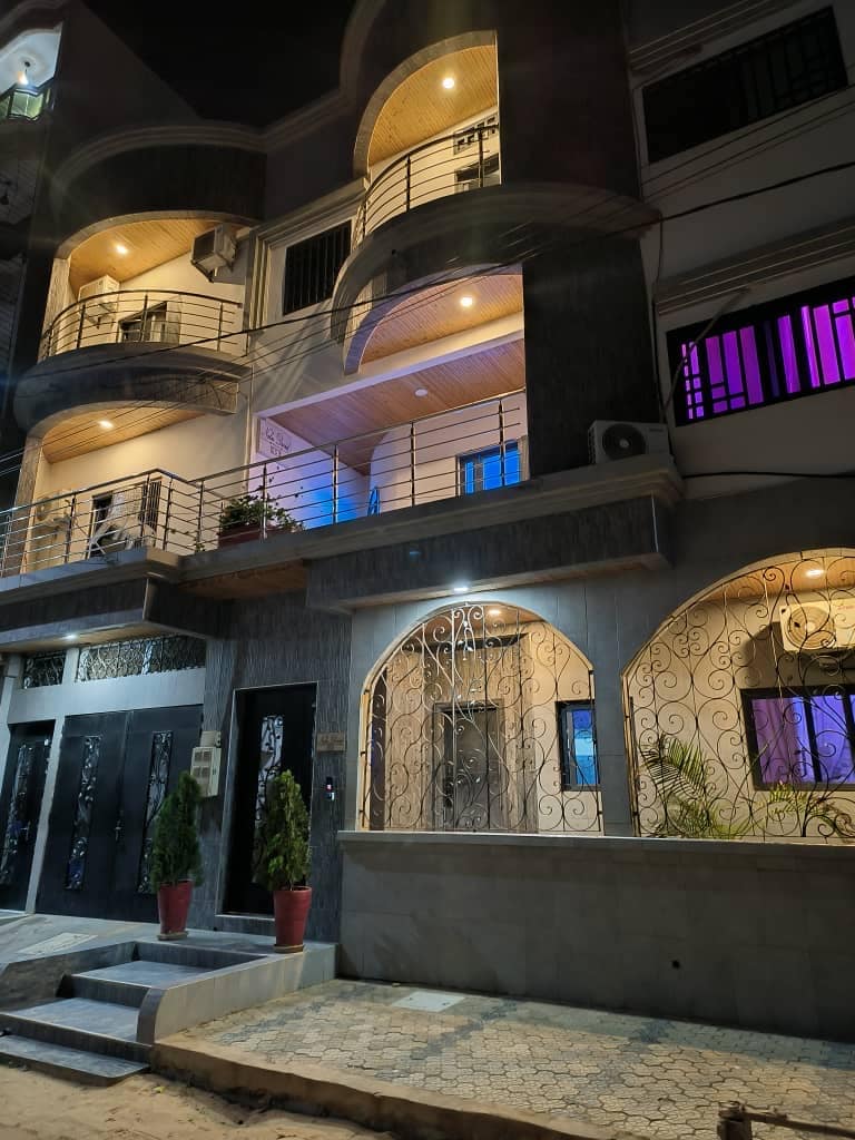 An elegant building is shown at night, featuring multiple balconies illuminated by soft lighting. Decorative architecture is highlighted, with wrought iron accents on doors and railings. Potted plants are positioned at the entrance, enhancing the welcoming appearance of the property.