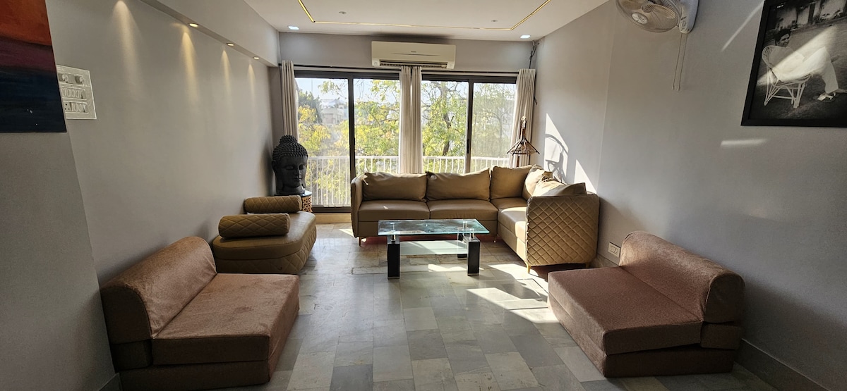 The living area features a light-filled space with a full glass wall providing views of greenery outside. Comfortable seating is arranged around a central glass table, with a mix of sofas and lounge chairs. Soft tones and natural light create a welcoming environment.