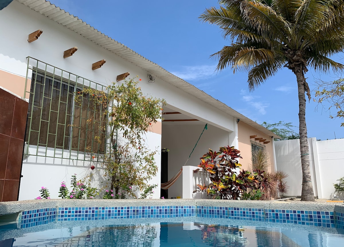 The exterior area is highlighted by a swimming pool bordered by colorful plants and flowers. A palm tree stands tall nearby, with a white-walled house featuring large windows and a sloped roof visible in the background against a clear blue sky.