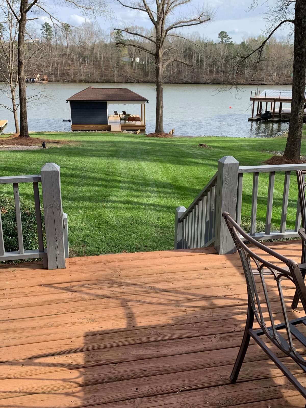 A view from the deck showcases well-maintained green grass leading down to the water's edge. A boathouse is visible on the opposite shore, with wooden seating and a dock extending into the lake. Sunlight gently illuminates the serene outdoor space.