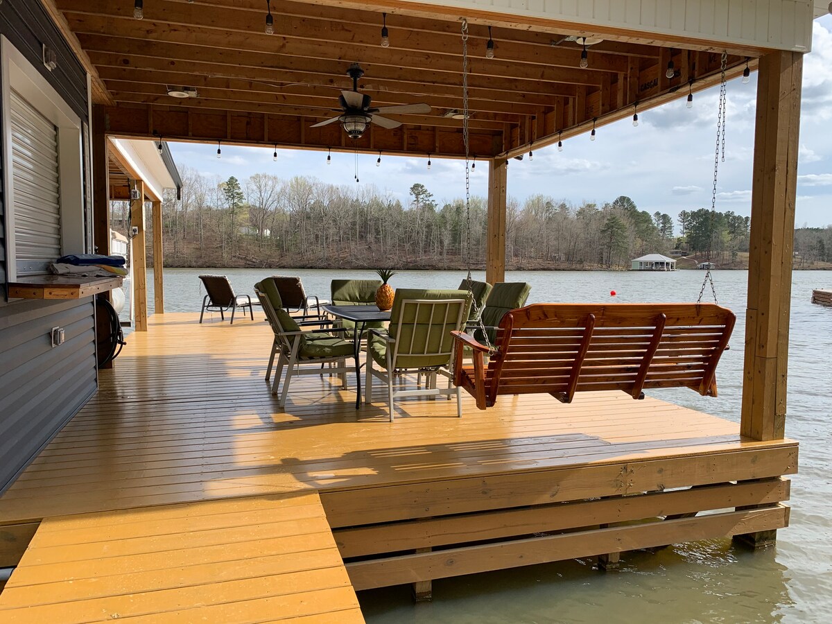 An inviting boathouse deck is visible, featuring a swing and a dining table surrounded by six green chairs. A ceiling fan provides airflow, while the serene lake and surrounding trees are seen in the background. The space is illuminated by hanging lights, enhancing its charm.