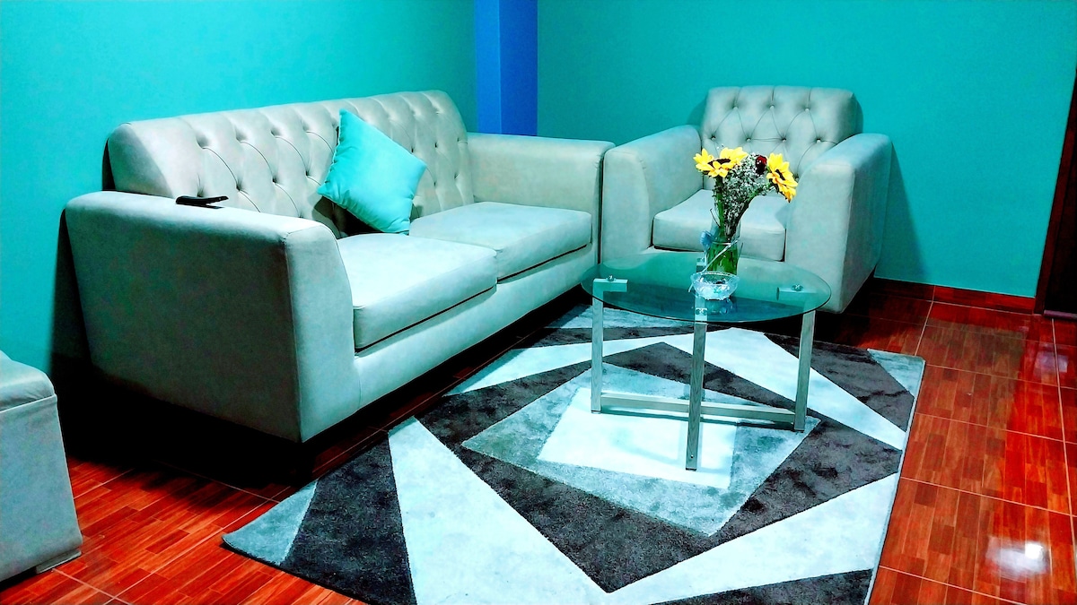 A cozy living area is highlighted by a light-colored sofa and armchairs, situated around a glass coffee table. A geometric rug lies on the polished floor, and a vase with fresh flowers adds a touch of color to the space. The walls are painted in a calming teal.