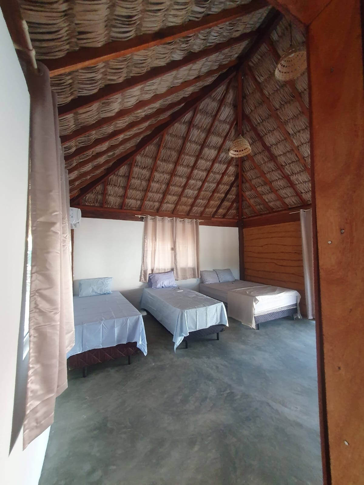 A spacious room features a high thatched ceiling with wooden beams. Three beds, each covered with light-colored linens, are arranged thoughtfully. Natural light enters through the large windows, and soft curtains drape elegantly beside them.