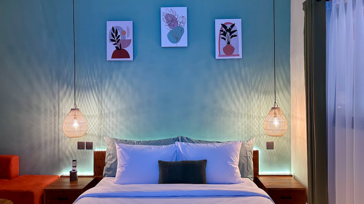 A serene bedroom features a spacious bed dressed in crisp white linens. Decorative wall art hangs above, complemented by woven pendant lights on either side. Soft blue-green walls create a calming atmosphere, while gentle lighting adds warmth to the space.