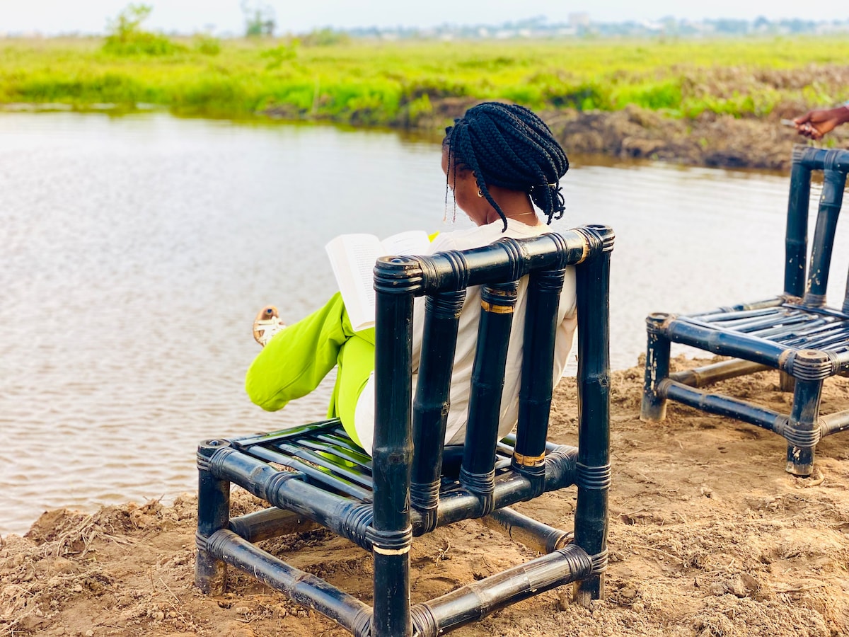 Two bamboo chairs are positioned on the edge of a tranquil body of water. A figure is seated in one chair, engaged in reading a book, overlooking the serene landscape. Lush greenery stretches beyond the water's edge, creating a peaceful natural setting.