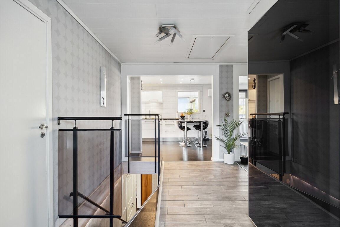 A hallway is displayed with sleek, light-colored walls and contrasting dark accents. The space is accentuated by a modern railing and a potted plant. Light wooden flooring leads to an open area, visible in the background, featuring a dining table and kitchen.