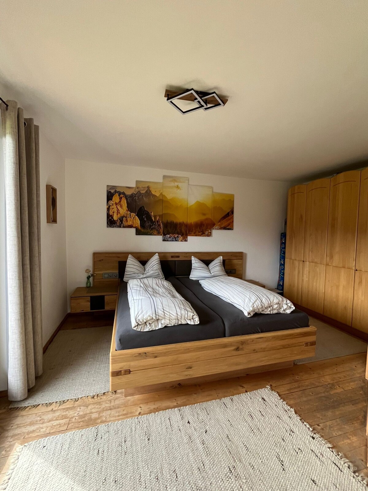 A cozy bedroom features a wooden double bed with striped bedding, accompanied by two bedside tables. A large wardrobe provides ample storage space. Natural light filters through the window, illuminating the mountain-themed artwork on the wall, while a neutral rug adds warmth to the wooden floor.