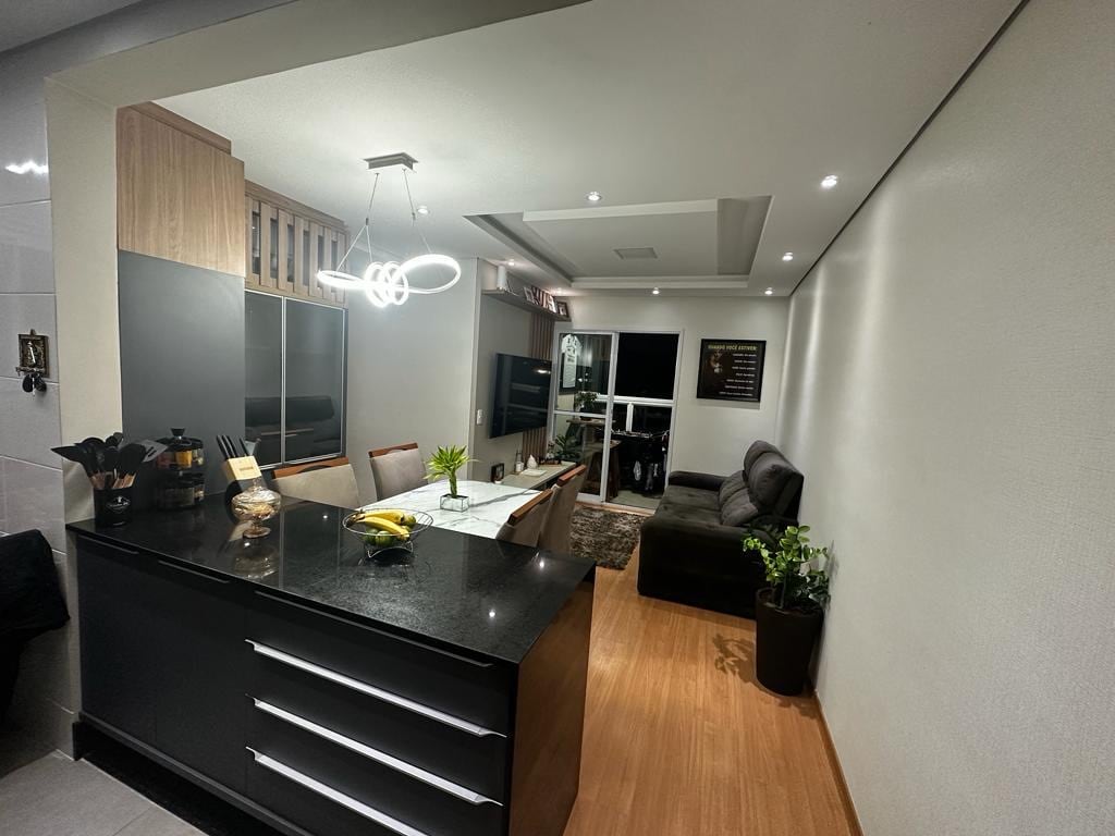 A modern kitchen and dining area feature sleek black cabinetry and a central island. A stylish light fixture hangs over the dining table, surrounded by wooden chairs. A comfortable seating area with a dark sofa is visible, complemented by decorative plants and warm wooden flooring.