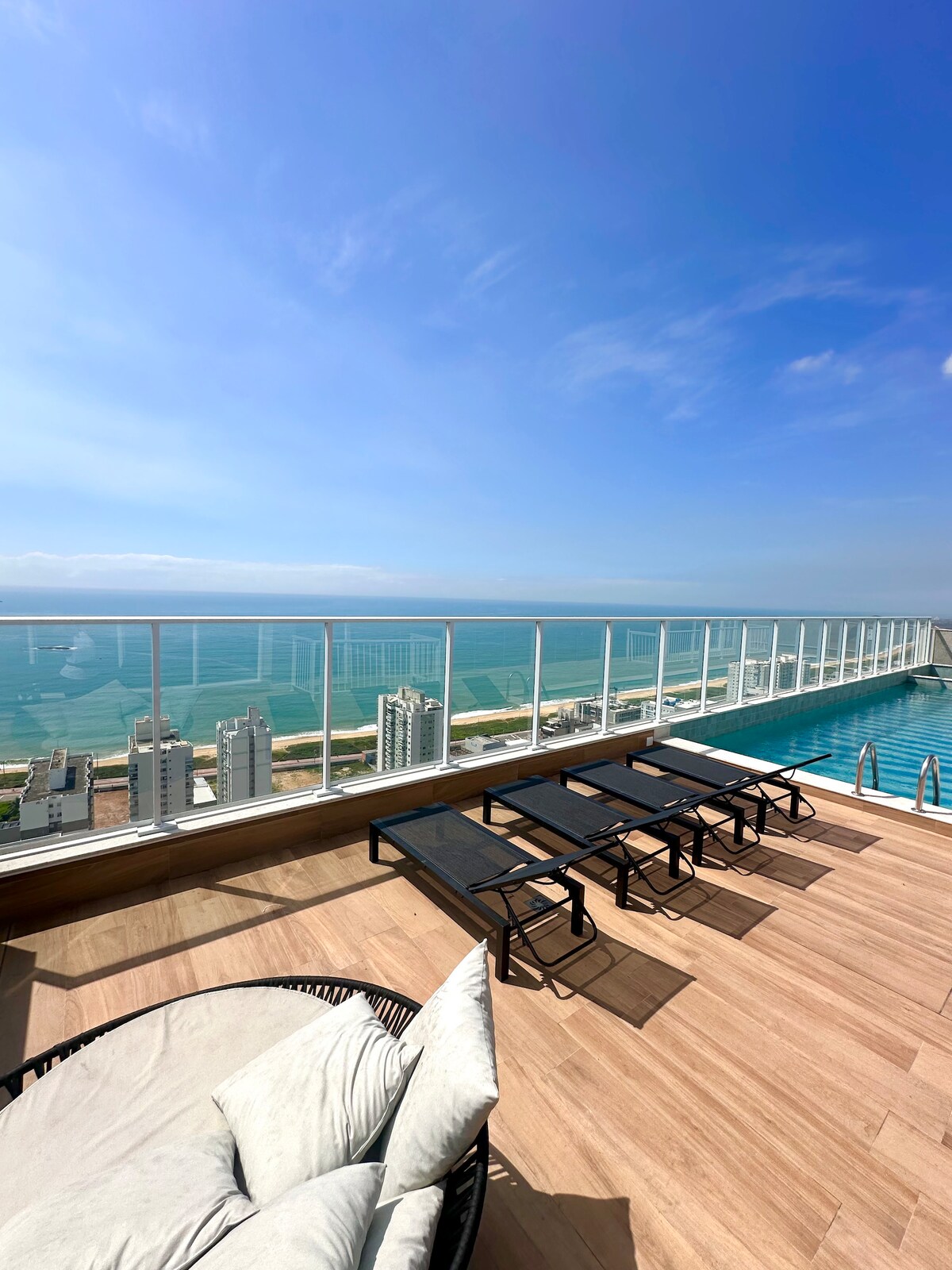 An outdoor terrace offers stunning views of the ocean and skyline, featuring a sparkling pool and multiple lounge chairs positioned for sunbathing. A round, cushioned seating area provides additional comfort, while the expansive glass railing enhances the openness of the space against a clear blue sky.
