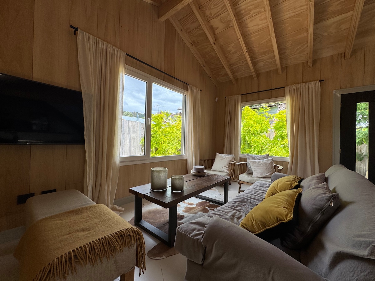 A bright living area is designed with natural wood accents and features large windows showcasing greenery outside. A comfortable couch is paired with decorative cushions, while a coffee table holds candles and a few simple decor pieces, creating a serene environment for relaxation.