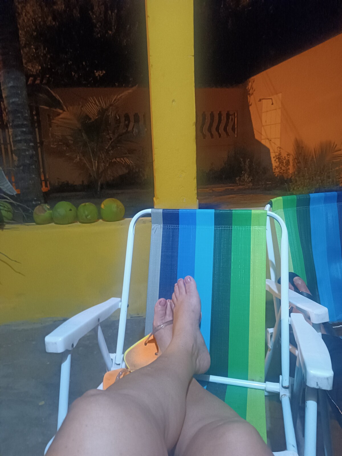 A pair of beach chairs with colorful striped patterns is seen in a relaxed outdoor area. The feet of a person are resting on one chair, with a backdrop featuring a vibrant yellow wall and lush greenery. Soft evening lighting enhances the tranquil ambiance.