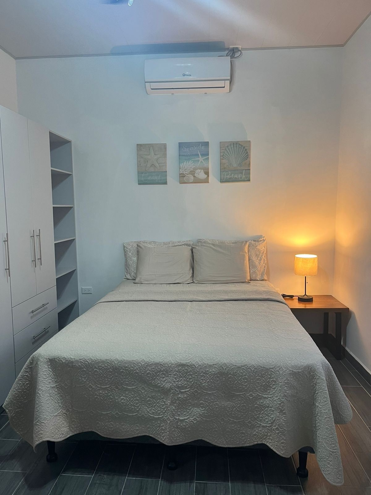 A simple bedroom is enhanced by soft lighting from a bedside lamp. The spacious bed is covered with a light-colored bedspread, and wall decor featuring beach themes is displayed above. A wardrobe and shelves are present, providing storage options.