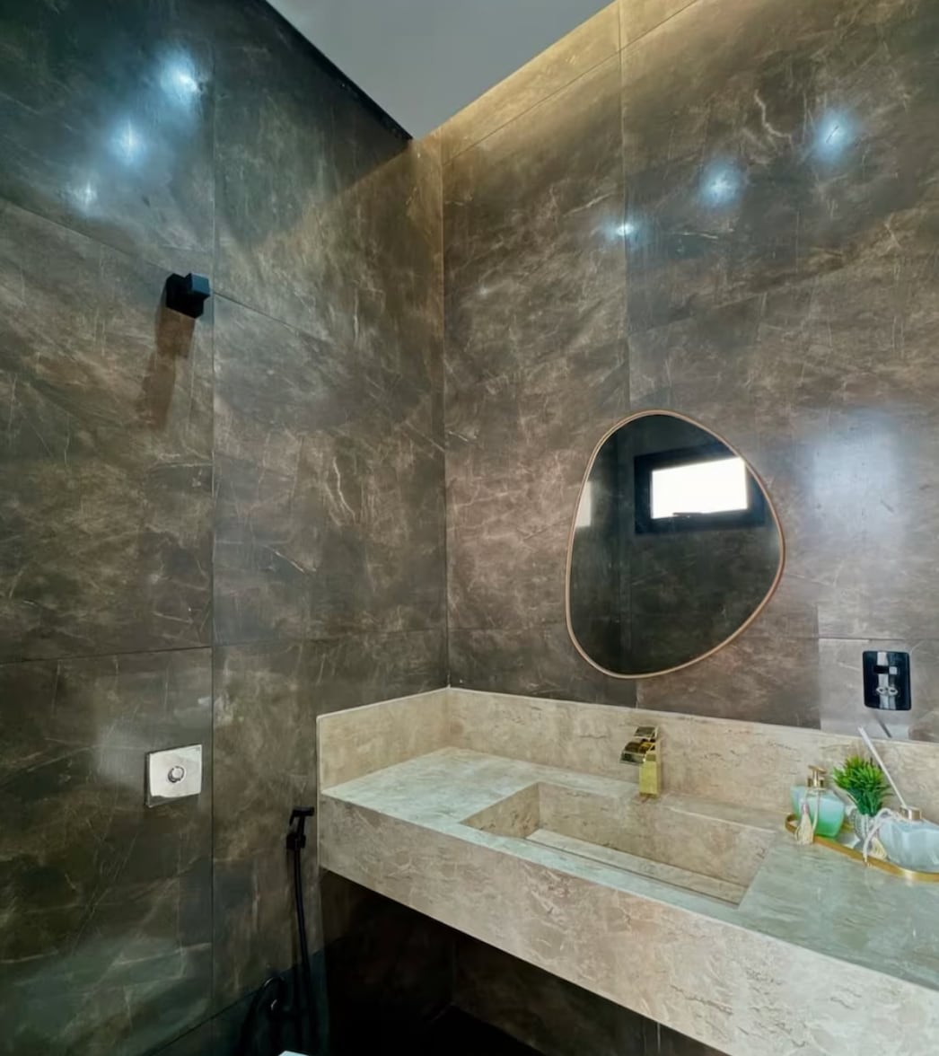 The bathroom features sleek, dark marble walls that create a modern look. A stylish oval mirror is mounted above a minimalistic sink with a gold faucet. Natural light streams in from a small window, enhancing the elegant design of the space.