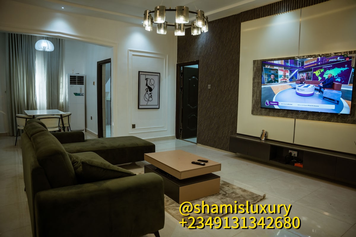 A modern living area features a large, plush green sofa facing a sleek wall-mounted television. A stylish coffee table sits in front, complementing a light-colored stone floor. Soft lighting from a contemporary ceiling fixture enhances the space, while a dining area is visible in the background.