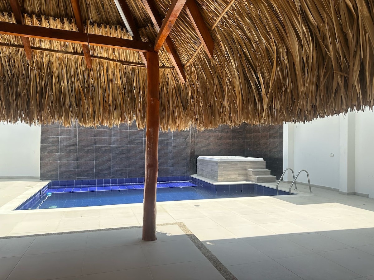 A shaded area under a thatched roof provides a view of a tranquil pool, featuring a light blue tiled surface. Surrounding the pool, smooth tiles lead to a deck area, ideal for relaxation and gatherings.