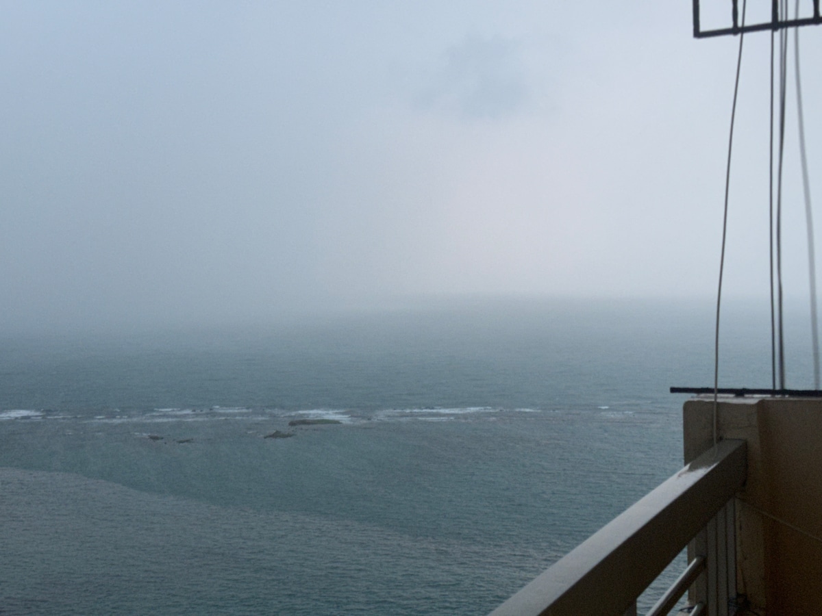 A panoramic view of the ocean is presented, featuring a vast expanse of water that blends into the horizon. The scene is shrouded in fog, creating a serene and muted atmosphere. A balcony railing is visible in the foreground, framing the view.