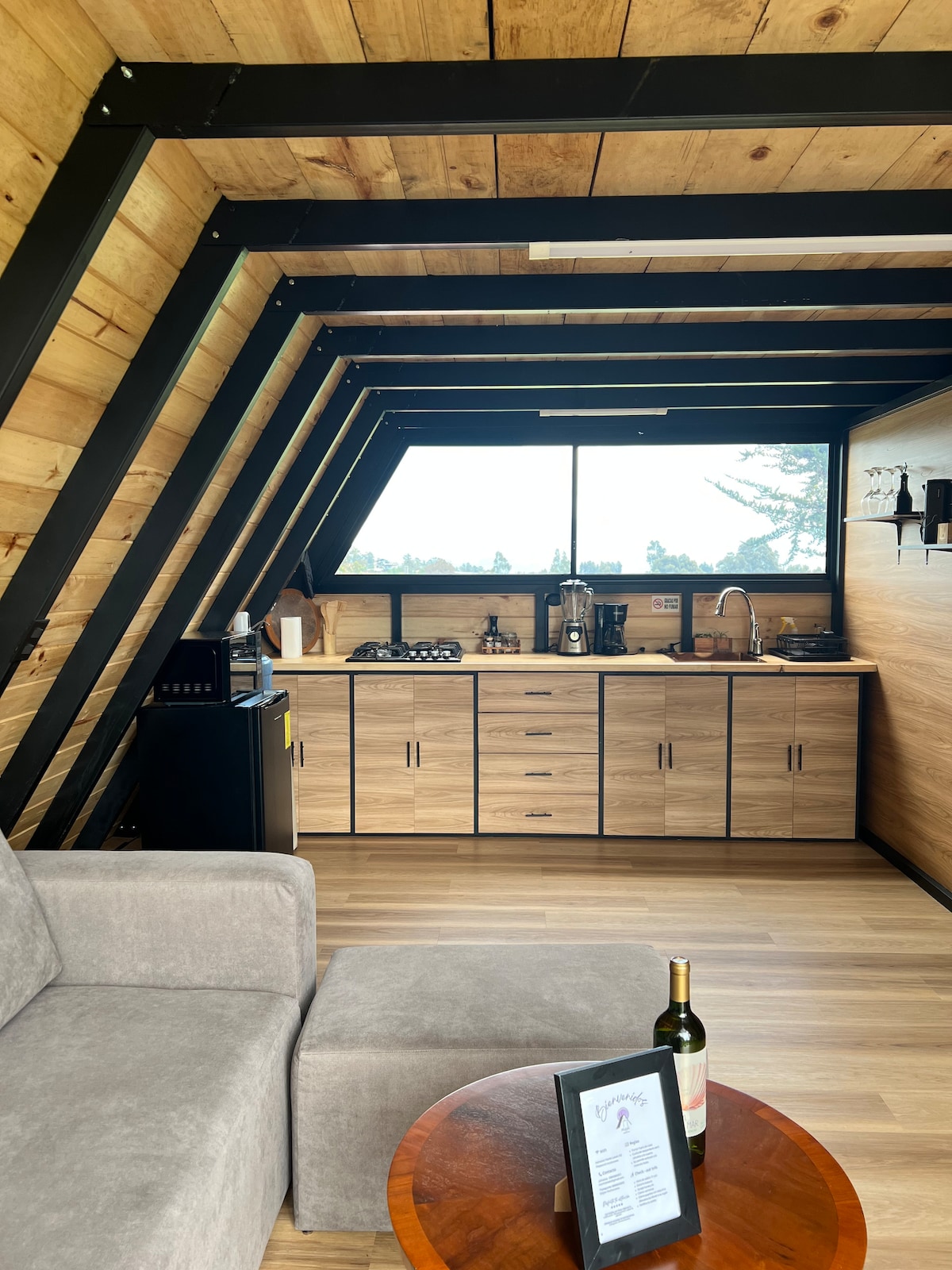 An A-frame cabin kitchen is shown, featuring wooden cabinetry and a countertop with a sink and stove. A small refrigerator is positioned nearby. The area is well-lit by a large window that offers a view of the surrounding landscape. A cozy seating area with a gray sofa and a round wooden table complements the space.