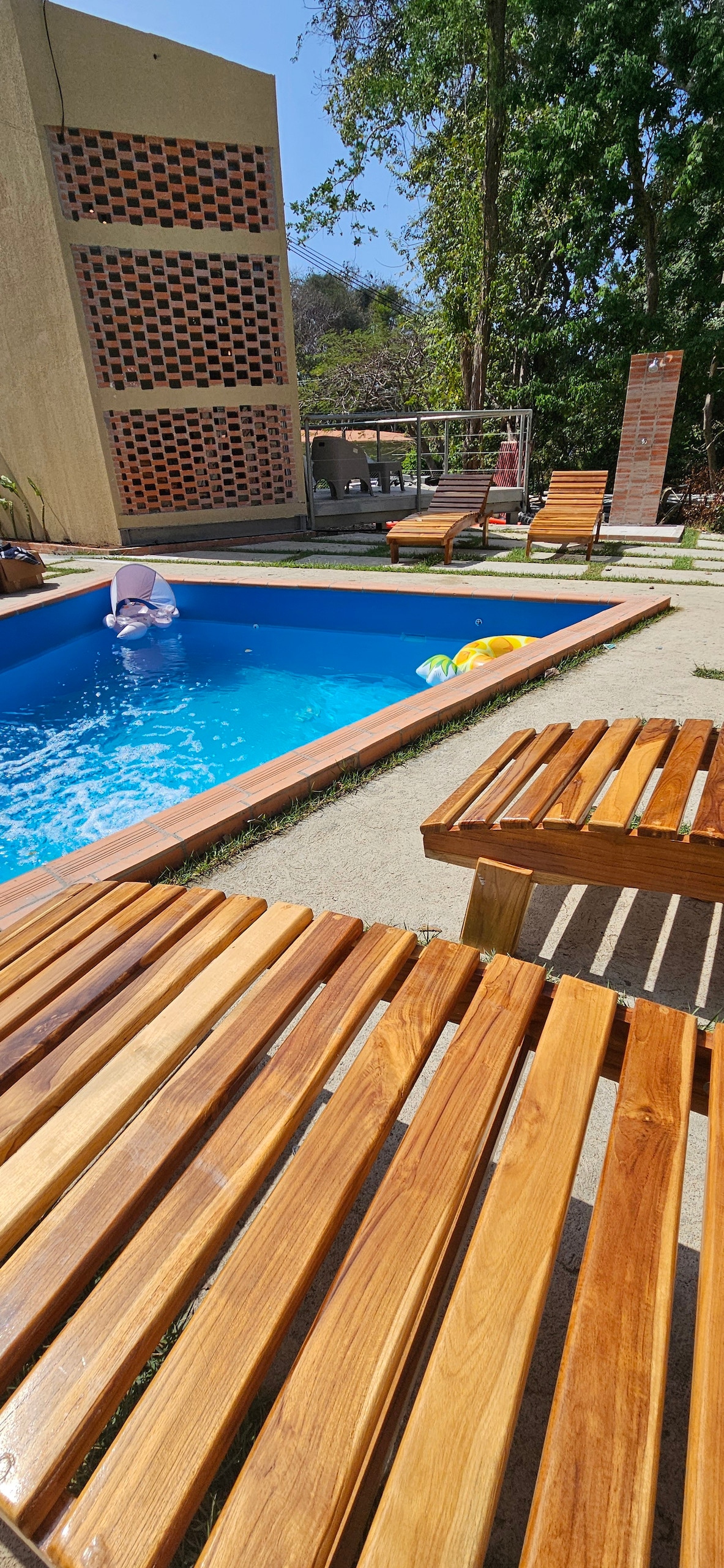 A small pool is set in a sunlit area, surrounded by wooden sunbeds. The pool water is clear, with colorful floats visible. A brick wall and outdoor kitchen area can be seen in the background, complemented by greenery providing shade.