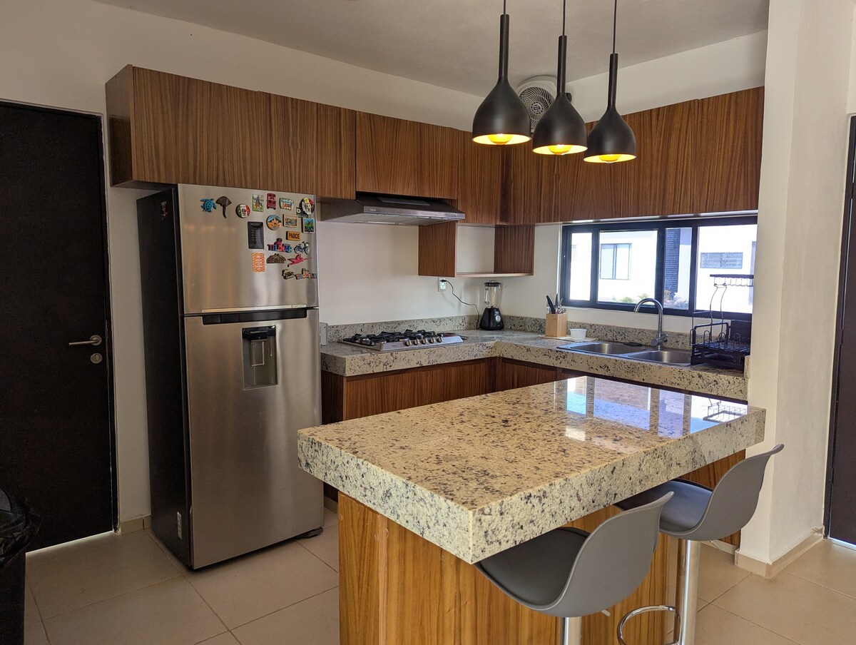 A modern kitchen features wooden cabinetry and a large granite island with two stools. A stainless steel refrigerator and a stove with an oven are visible, along with a coffee maker. Natural light enters through a window, illuminating the space.