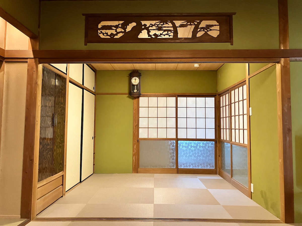 A serene traditional Japanese room is showcased, featuring tatami flooring and green walls. Sliding doors of varied textures create a harmonious atmosphere. A wooden clock can be seen on the wall, adding a touch of charm to the space illuminated by natural light.