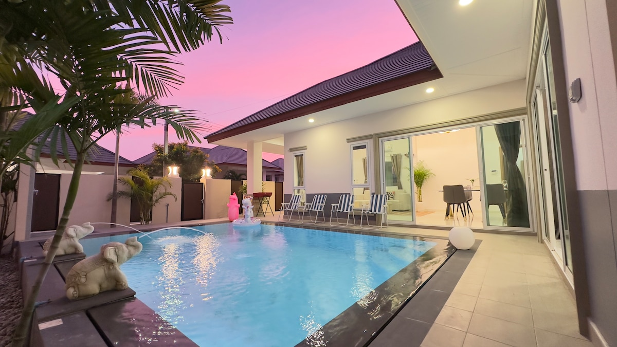 A private pool is set within a modern outdoor area featuring lounge chairs. The tranquil water reflects the soft colors of the evening sky. Nearby, a cozy living space can be glimpsed through large sliding glass doors.
