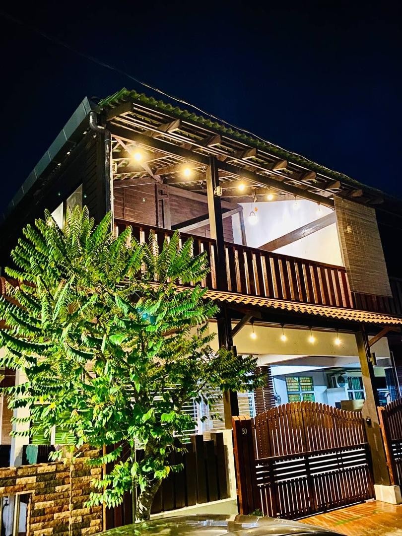 The two-story structure is illuminated by warm string lights, showcasing a balcony with wooden railings. A lush green tree is positioned in front of the entrance, enhancing the serene atmosphere. The building's exterior is constructed from a mix of wood and stone materials.