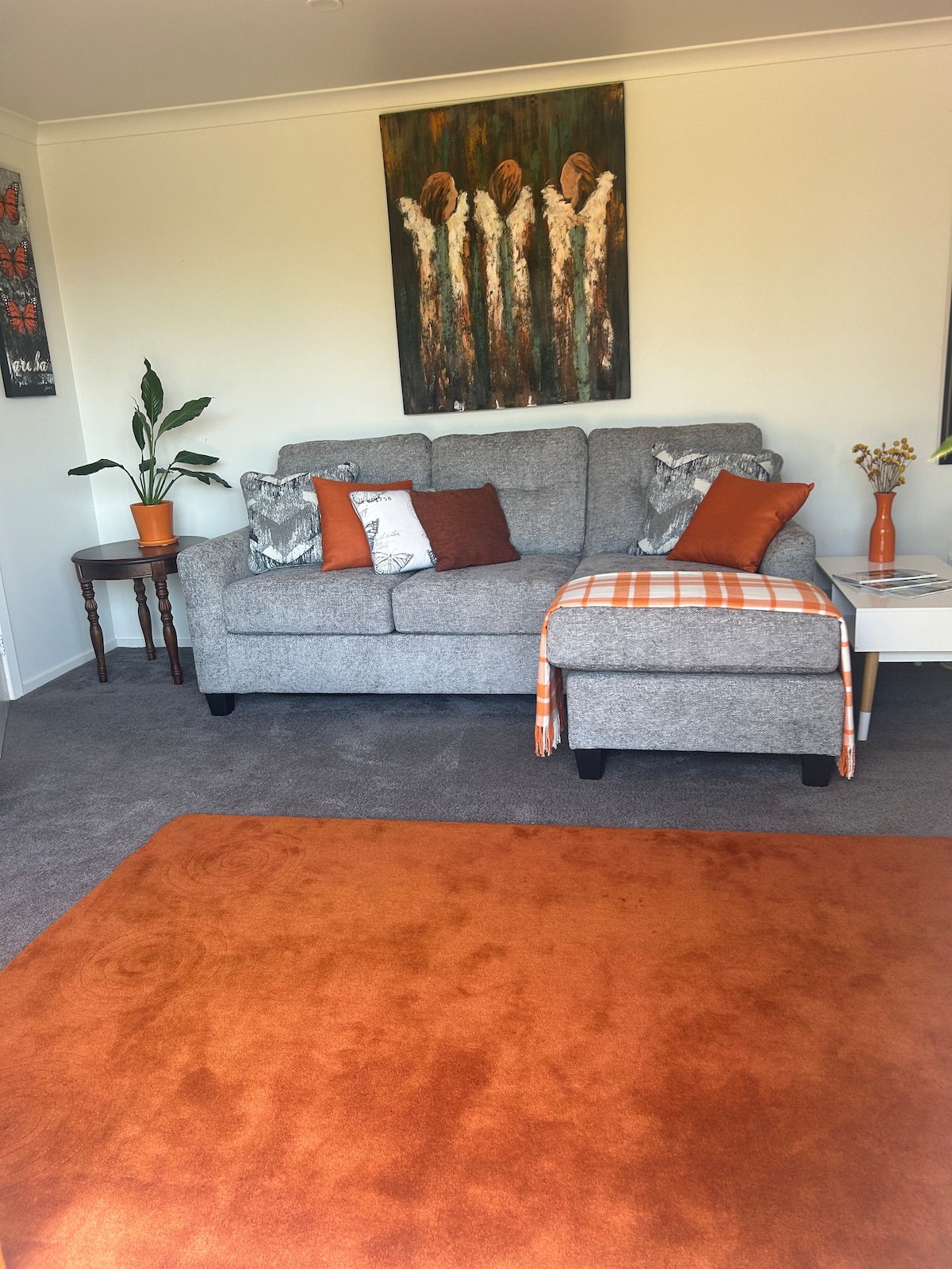 A comfortable seating area features a gray sofa adorned with colorful cushions and a warm throw. A small side table holds a vase with flowers, while a potted plant provides a touch of greenery. An orange area rug lies beneath, complementing the inviting decor.