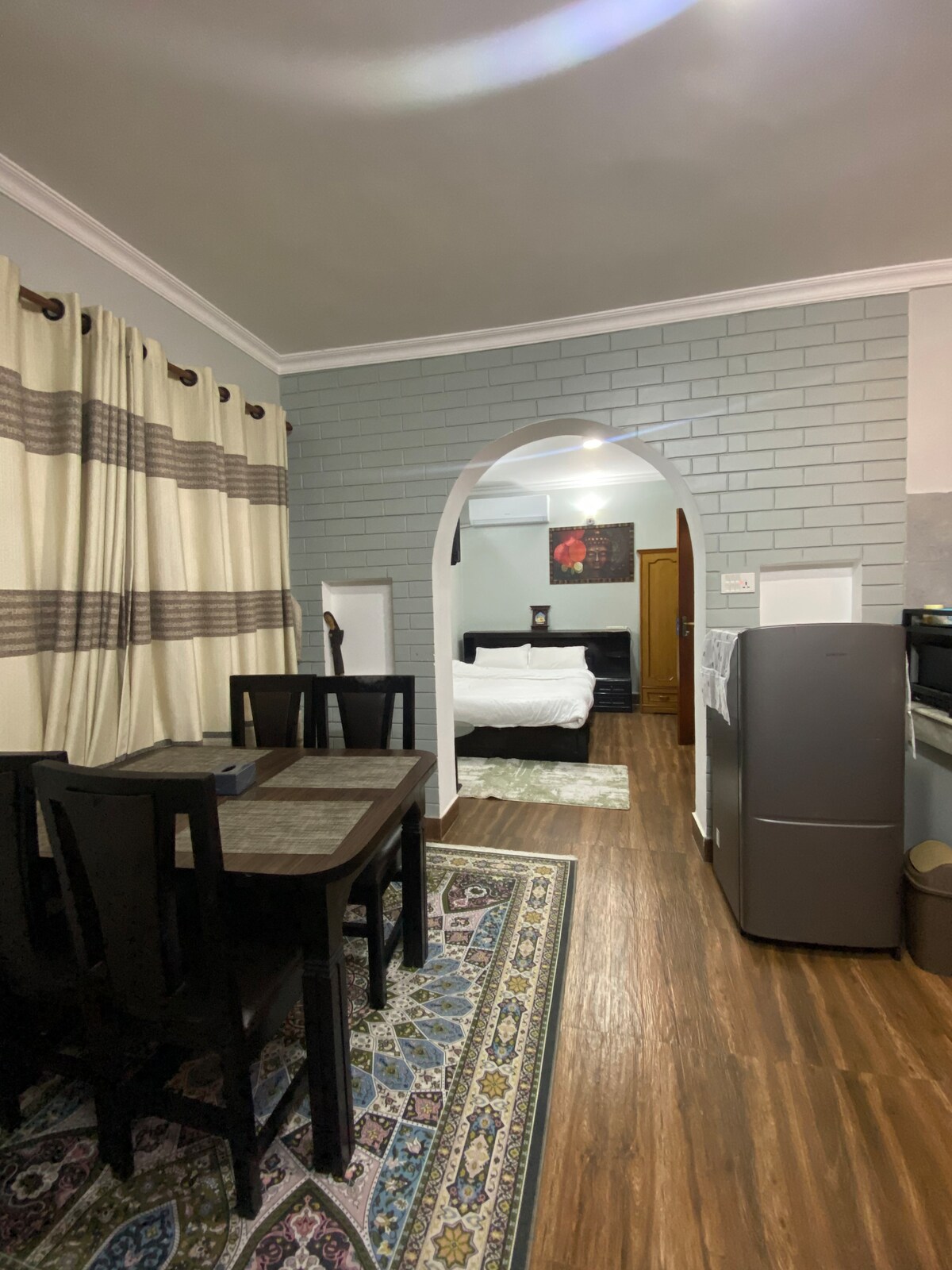 A dining area features a wooden table with four chairs, set against a patterned rug. Light-colored curtains with dark stripes frame a nearby window. An archway leads to a sleeping area, where a spacious bed and framed artwork are visible.
