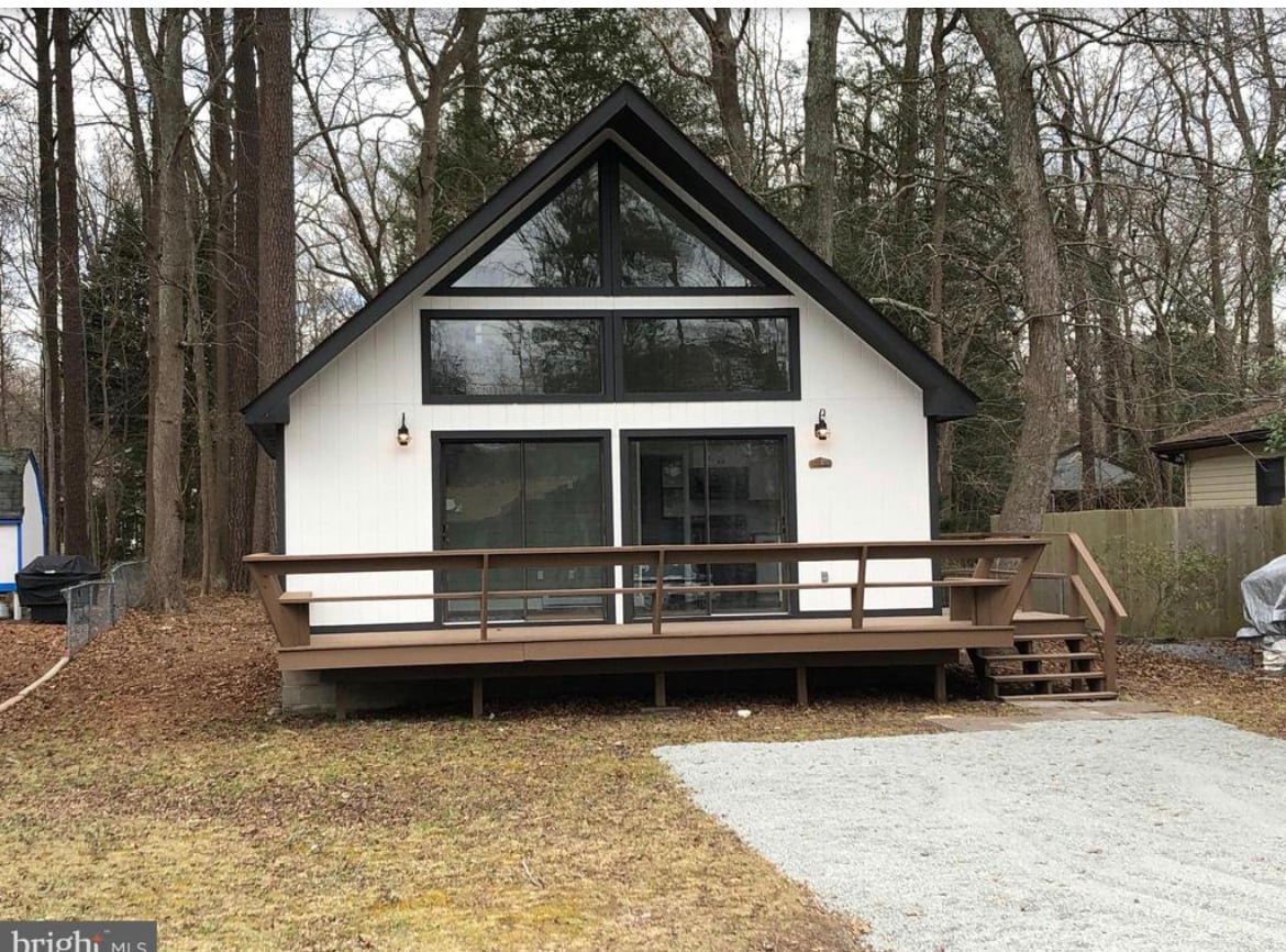 A modern cottage exterior is framed by tall trees, featuring a slanted roof and large glass windows that invite natural light. A spacious wooden deck with a railing extends from the front, providing an area for outdoor seating, surrounded by gravel and leafy ground.