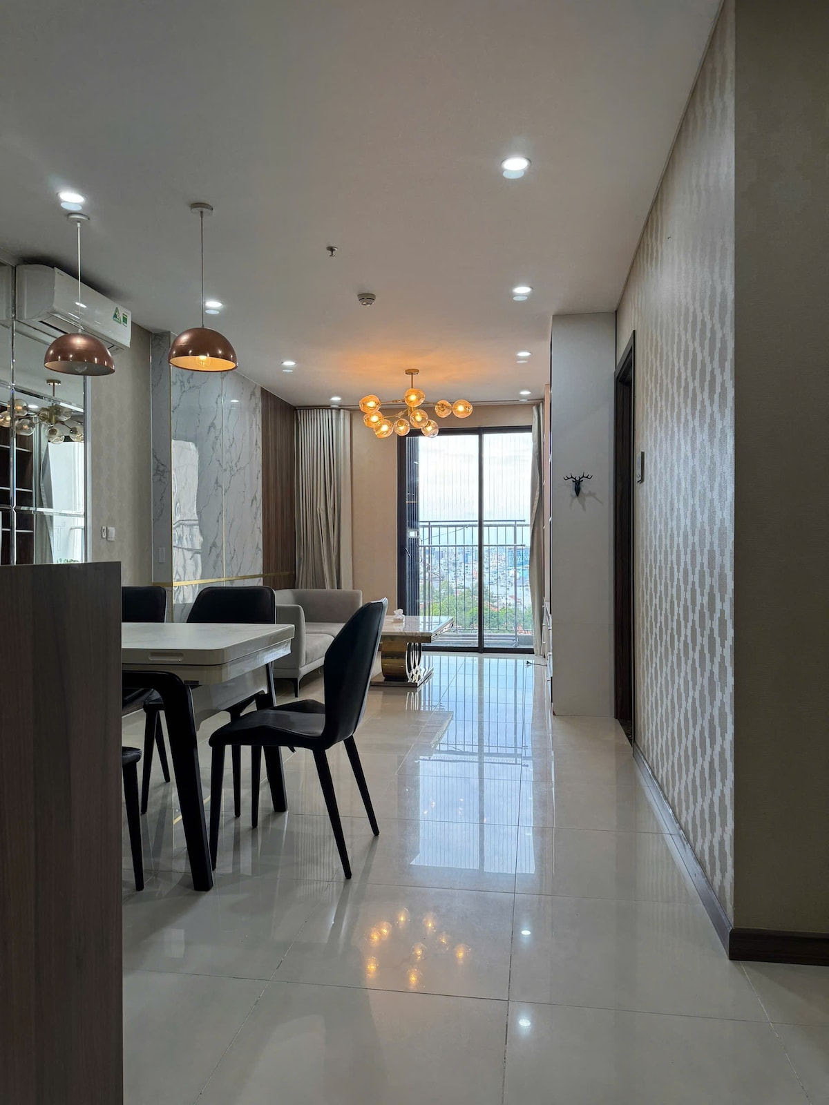 A modern dining area is highlighted by a rectangular table surrounded by black chairs. Soft lighting from ceiling fixtures enhances the spaciousness of the room. Large windows offer abundant natural light and a view of the cityscape beyond. Sleek tiled flooring completes the contemporary design.