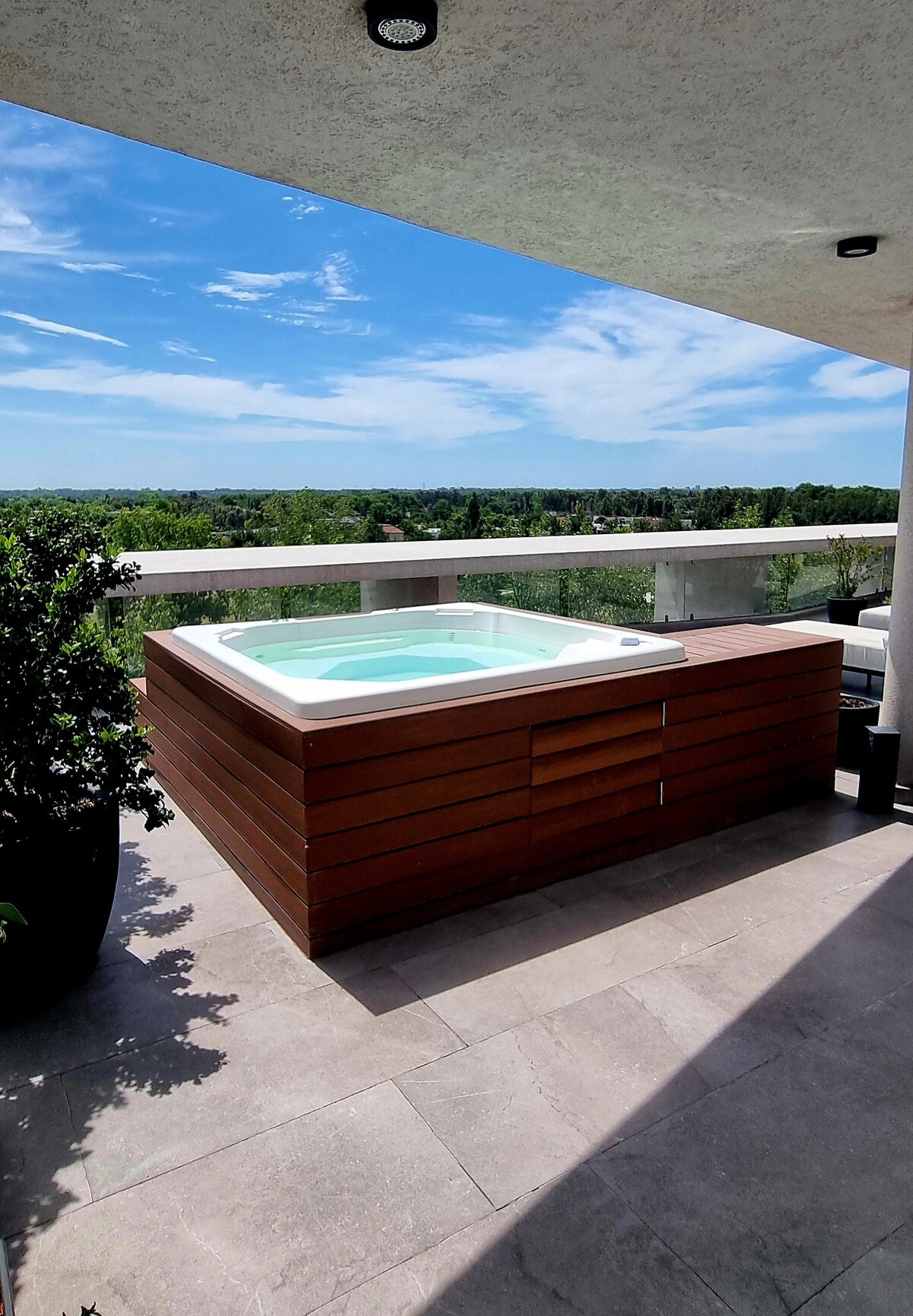 A wooden hot tub is positioned on a balcony, surrounded by potted plants. The serene landscape of greenery is visible in the distance under a bright blue sky. The floor features light-colored tiles, enhancing the outdoor space.