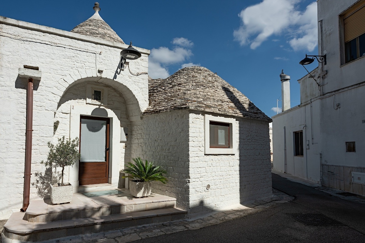 The exterior of a traditional stone structure is displayed, featuring a rounded roof of overlapping stones. The entrance is marked by a wooden door and a small step, with a potted plant positioned nearby. Clear blue skies create a serene backdrop against the light-colored building.