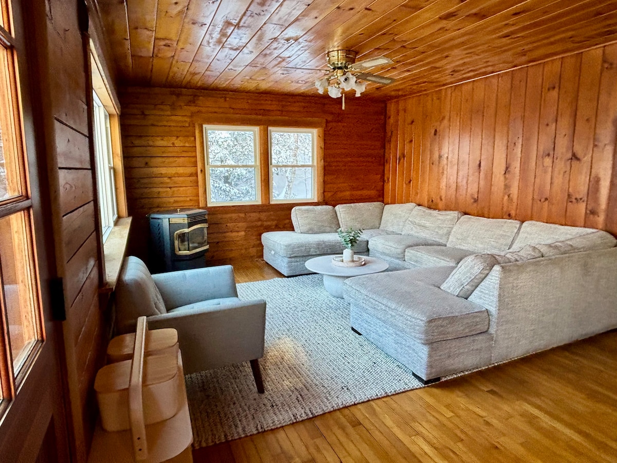 A warm living area features wooden walls and a light wood floor. A spacious sectional sofa is positioned around a circular coffee table, providing comfort for gatherings. Two large windows allow natural light to brighten the space, while a cozy chair adds an extra seating option.