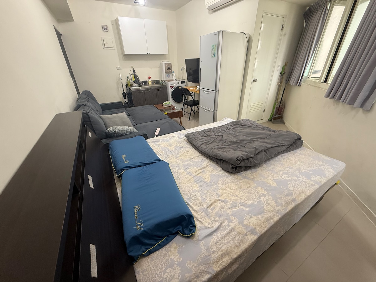 The living space features a double bed with a light cover, accompanied by two blue pillows. A gray sofa is situated beside the bed, and a dining table with chairs is positioned in the background. Natural light is provided by a window dressed with light-colored curtains.